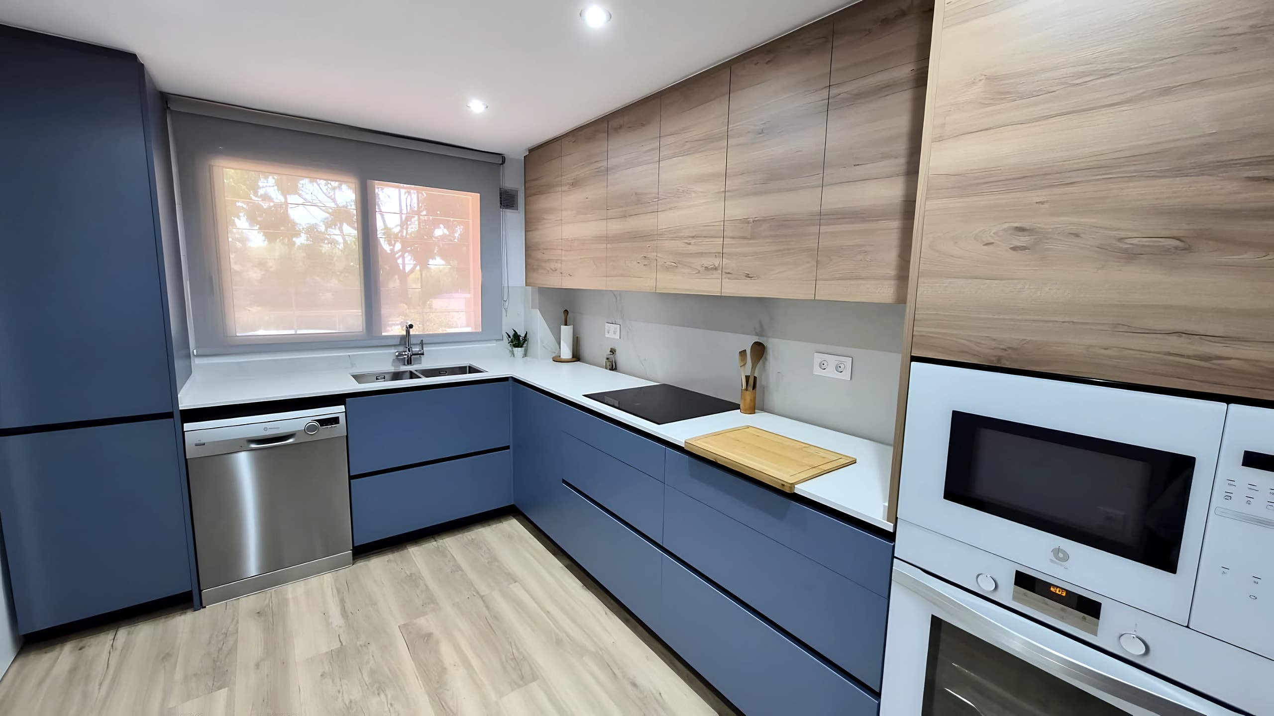 Cocina en U con frentes azules mate, altos en acabado roble, encimera blanca y horno y microondas blancos