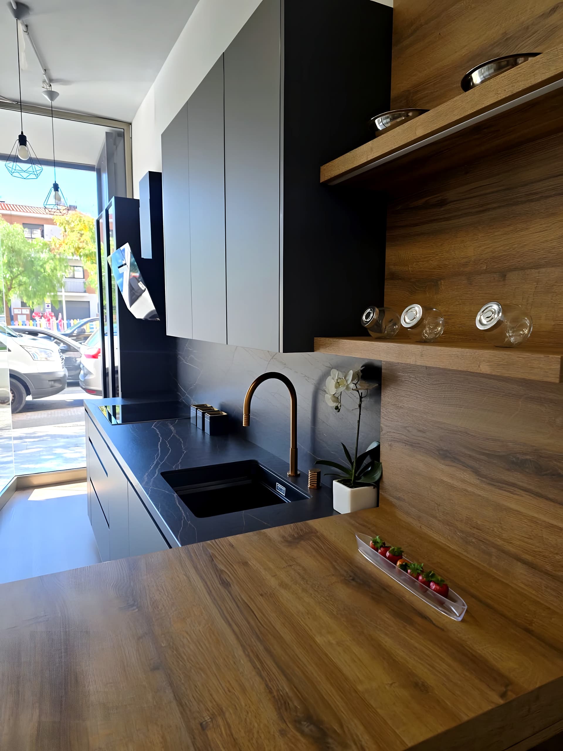 Fregadero negro bajo encimera oscura con grifo cobrizo junto a un panel de roble con baldas