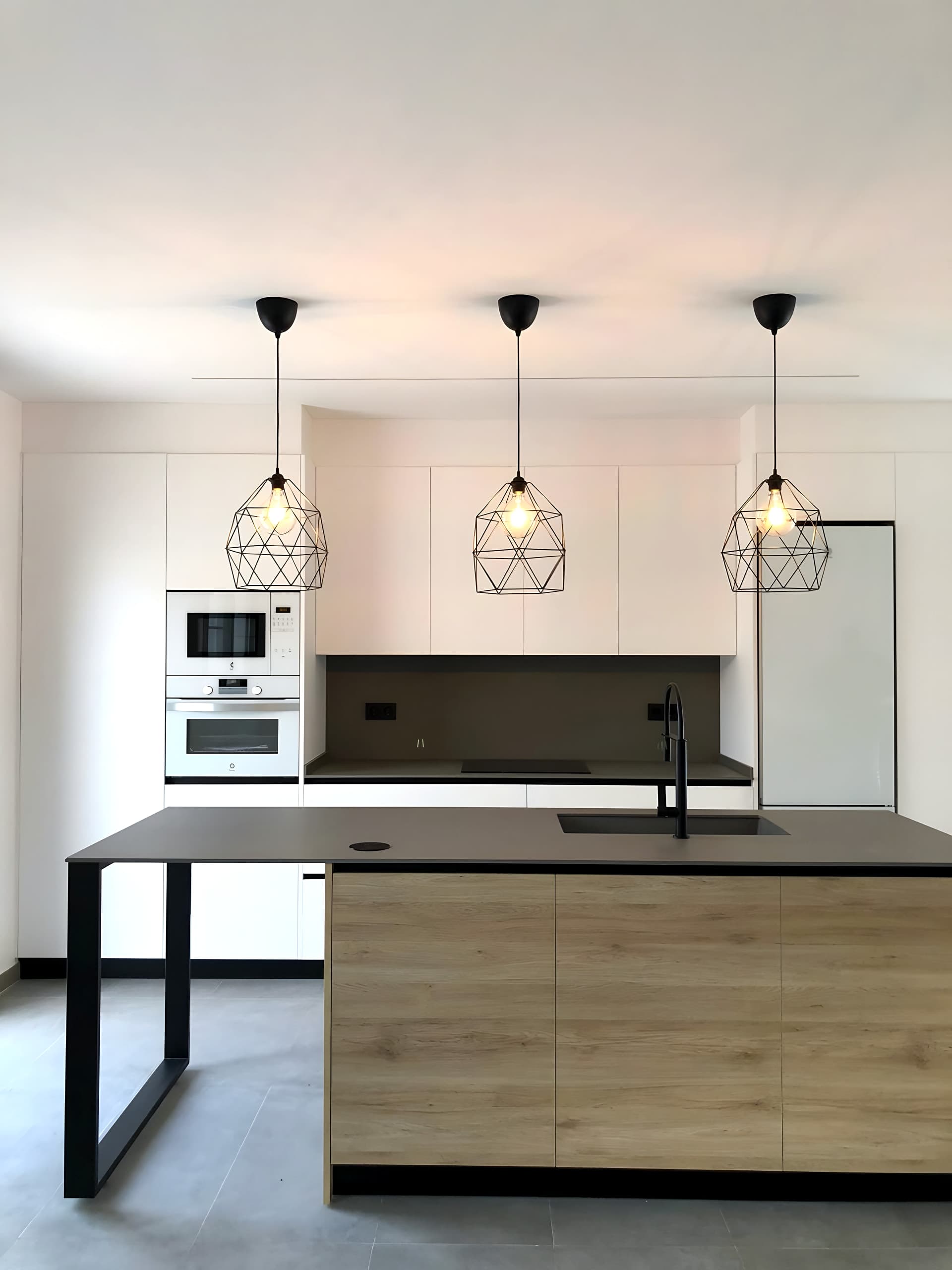 Cocina blanca con isla de frente en roble, encimera oscura, pata de acero negro y tres lámparas geométricas