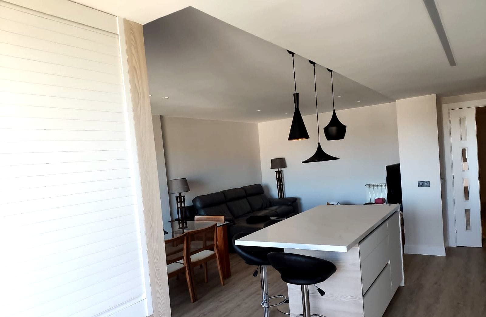 Cocina blanca abierta al salón con isla y taburetes bajo tres lámparas negras