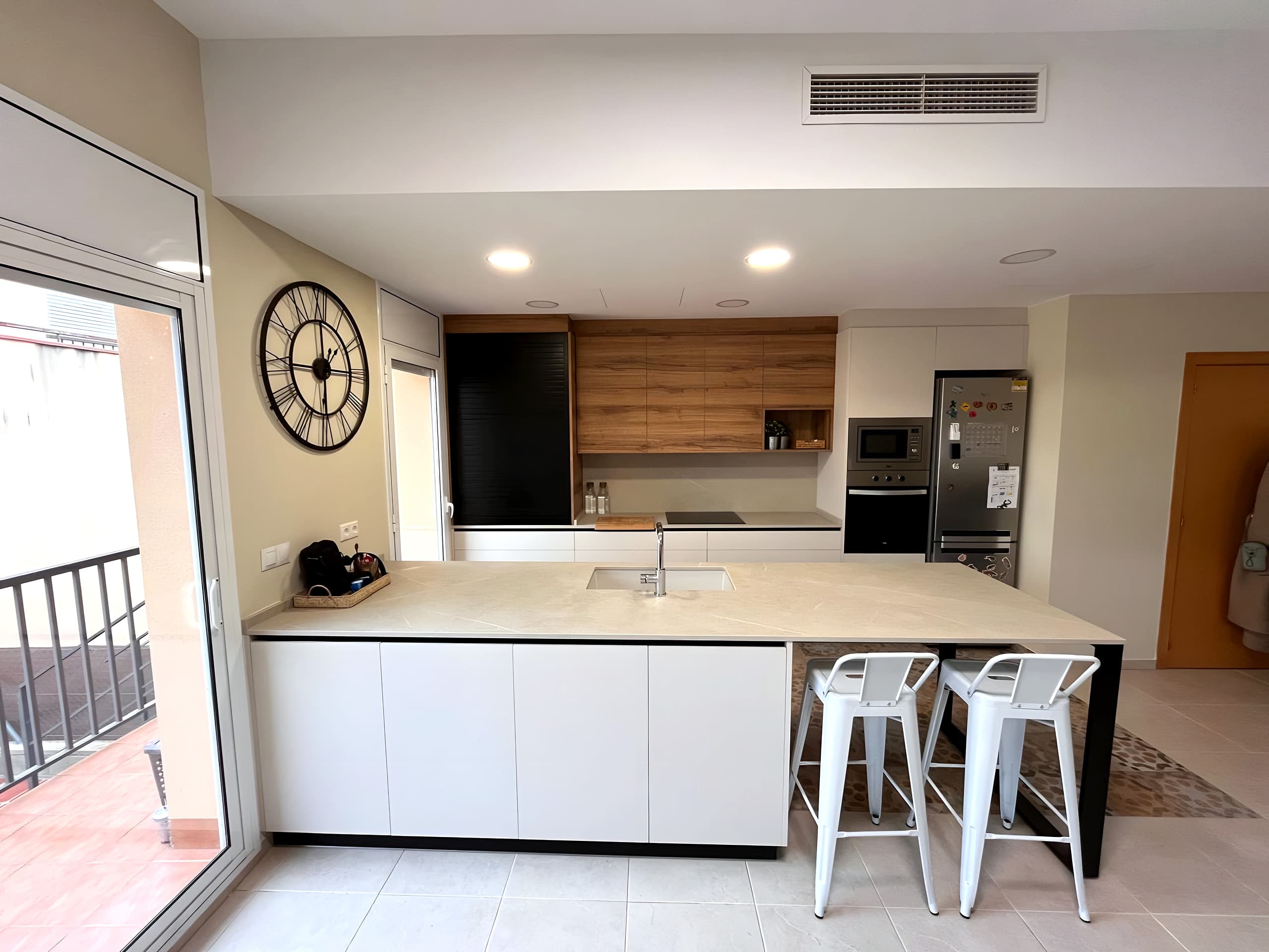 Isla blanca con encimera clara y barra sobre estructura negra, frente de cocina con muebles altos de madera