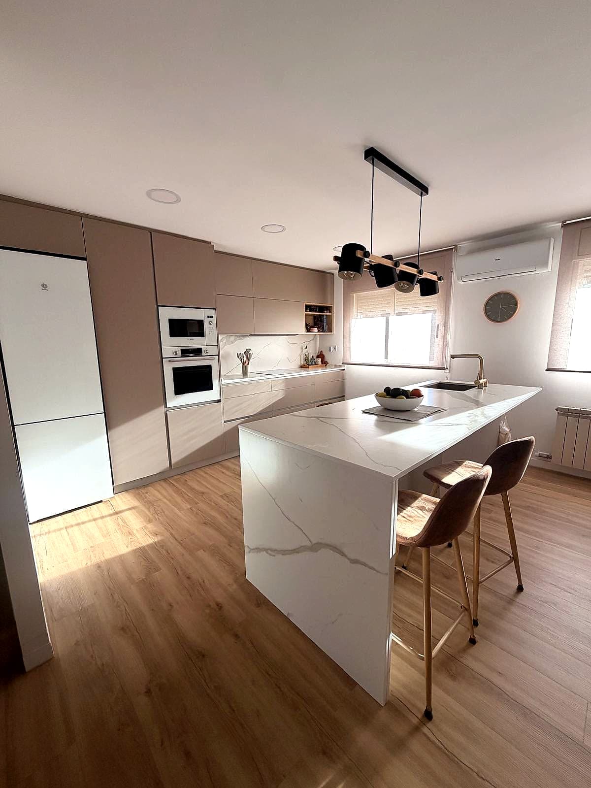 Cocina con columna de hornos blancos, frentes topo e isla blanca con taburetes junto a la ventana