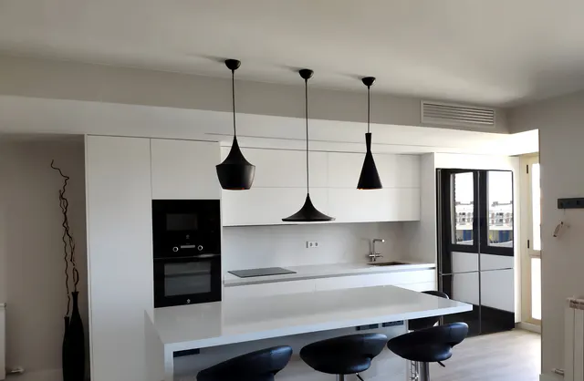 Cocina blanca con isla en cascada, barra con tres taburetes negros y tres lámparas colgantes negras