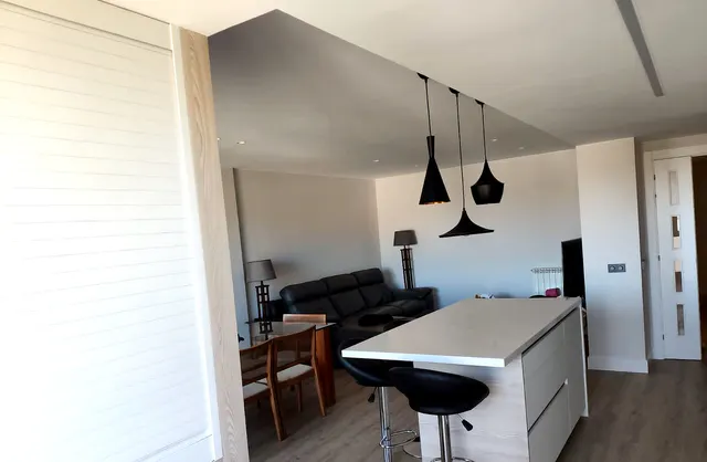 Cocina blanca abierta al salón con isla y taburetes bajo tres lámparas negras
