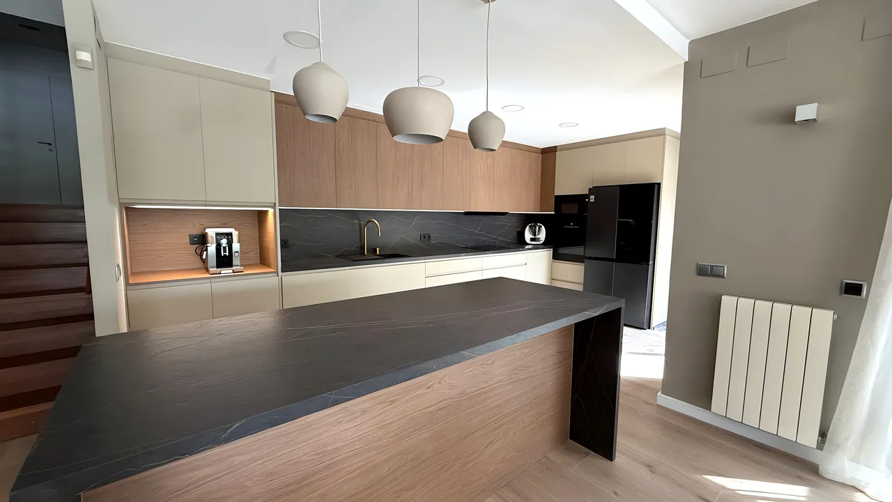 Cocina abierta con isla de encimera oscura veteada, muebles altos de madera, frentes crema y tres lámparas colgantes