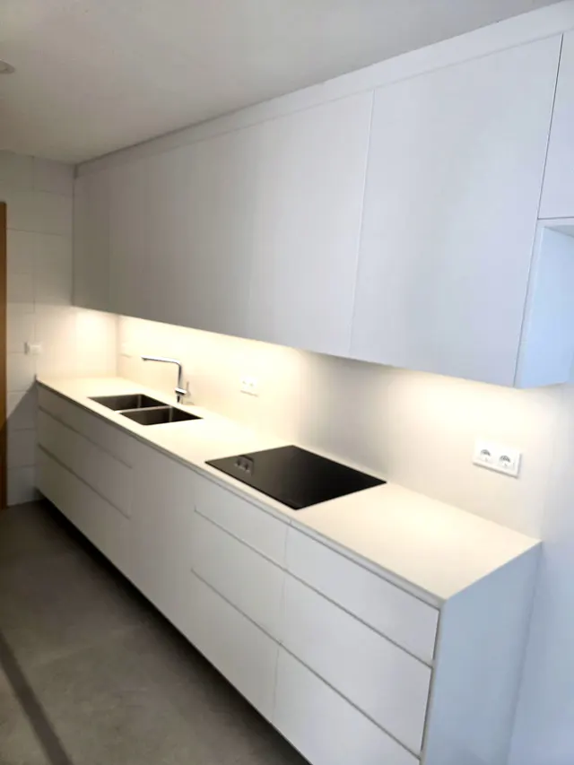 Frente blanco con fregadero doble, placa negra y muebles altos iluminados desde abajo