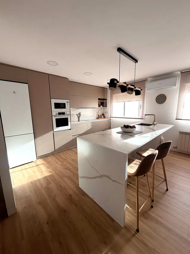 Cocina con columna de hornos blancos, frentes topo e isla blanca con taburetes junto a la ventana