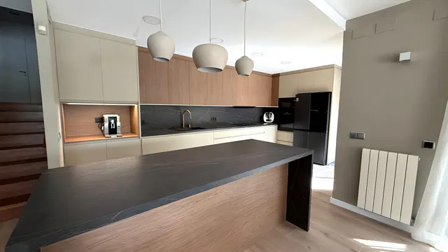 Cocina abierta con isla de encimera oscura veteada, muebles altos de madera, frentes crema y tres lámparas colgantes