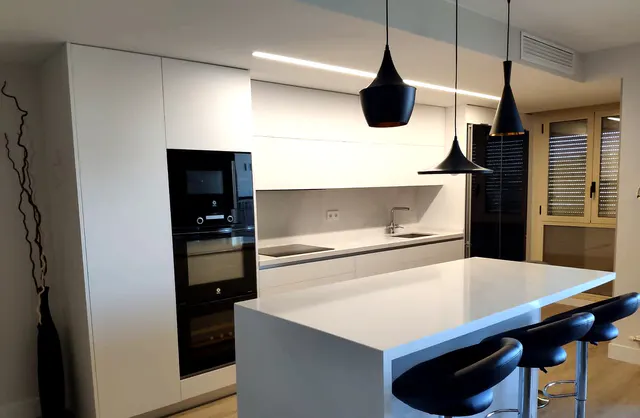Cocina blanca con columna de hornos negros e isla blanca con taburetes junto a la ventana